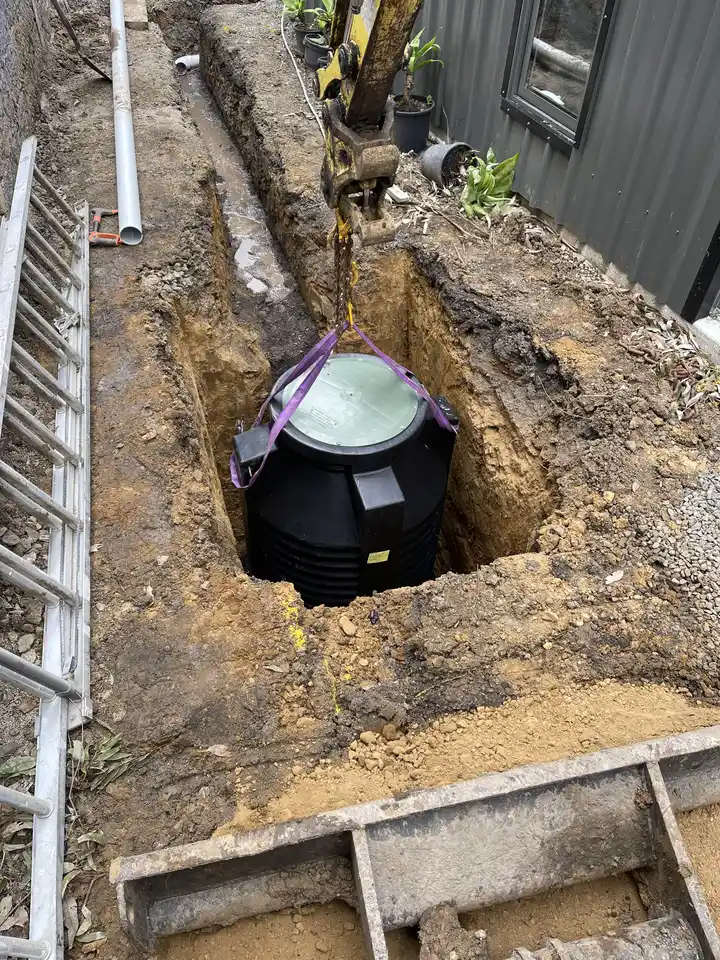 Excavator lowering a large underground water tank into a deep excavation hole