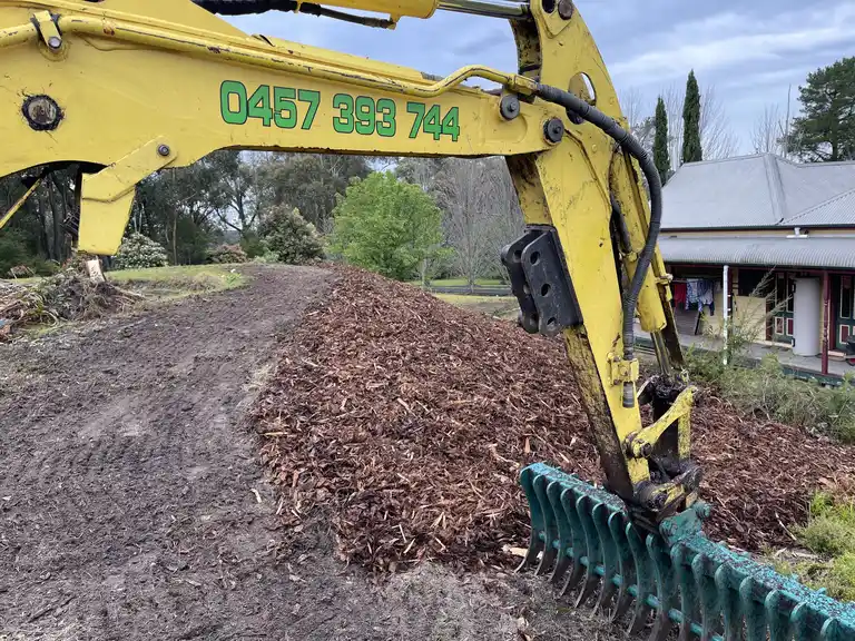 Excavator with rake attachment spreading mulch on a large sloped property